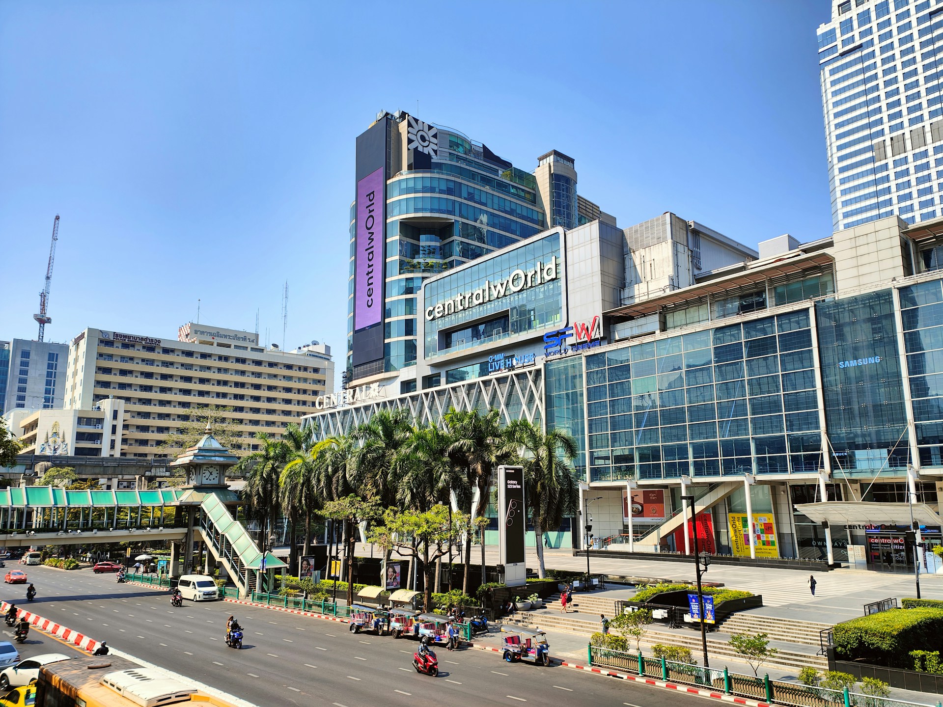 A modern office building with a "For Sale" sign out front, surrounded by lush landscaping and a bustling cityscape in the background