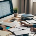 A freelance writer working on real estate articles at a cluttered desk with a computer, notebooks, and reference materials spread out