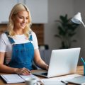 A blonde female content creator in dungarees working in her home office creating blog posts to generate leads for her home services client.