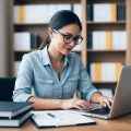 A female writer with glasses working on her laptop surrounded by folders and books about real estate case studies.