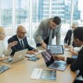 A group of real estate professionals collaborating around a table with digital devices and brochures in a bright office.