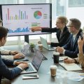 Business professionals collaborating around a conference table analyzing charts and graphs on laptops and a large digital screen.
