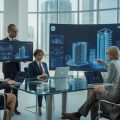 A group of professionals collaborating in a modern office with digital screens showing real estate data and 3D building models.