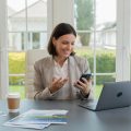 A real estate agent sitting at a desk using a laptop and smartphone in a bright home office with windows showing houses outside.