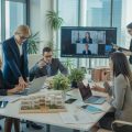 A group of professionals collaborating in a modern office with some working remotely via video calls and others meeting in person around a conference table with real estate materials.