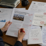 A professional editor meticulously compares a printed real estate article draft with various reference materials spread across a desk, including market data charts, formal property records with official stamps, and several pages of scribbled handwritten review notes. The scene captures the tactile details of the paper documents, the distinct ink of the annotations, and the focused atmosphere of a detailed fact-checking process in a well-lit workspace.