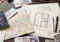 An organized arrangement of an international passport, detailed architectural floor plans, various paper currency notes, and a folded city map spread across a desk. The scene emphasizes the textures of the paper documents and the professional atmosphere of a global real estate review, with clear details on the blueprints and the map's markings.